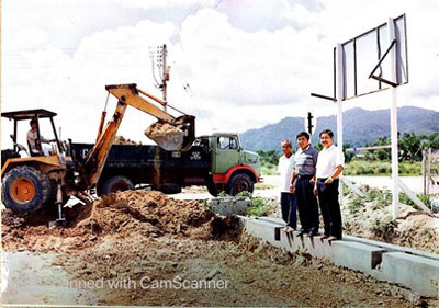 A photo shows workers operating an excavator and a load-carrying truck in a construction site. Three formally dressed men standing on a concrete slab beneath a hoarding on the right side of the photo are posing for the camera.