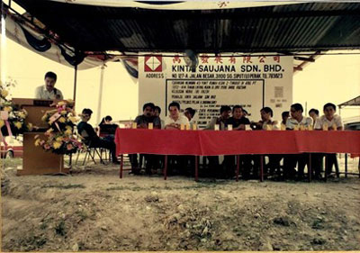 A man standing on a podium is speaking on the mike. A group of people dressed in formal attire are on the chairs beside the podium. A hoarding behind the people reads “Kinta Saujana SDN.BHD.