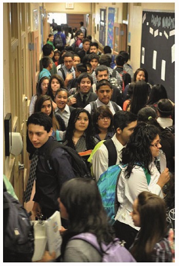 An image shows a narrow hallway crowded with students. Several bulletin boards and charts are stuck on the right wall of the hallway and few doors are on left side.
