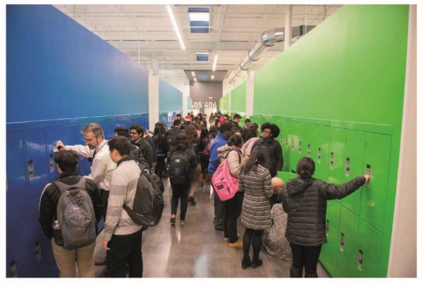 Image shows a high school hallway crowded with students. Students are using the lockers and racks on either side of the hallway. Few teachers are guiding the students. Label “05406” is on one wall across the hallway.