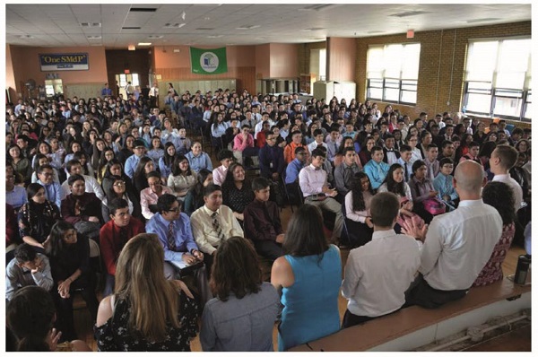 Three men and four women are addressing hundreds of high school students seated before them in a room. A sticker on the wall behind reads “One SMdP.”