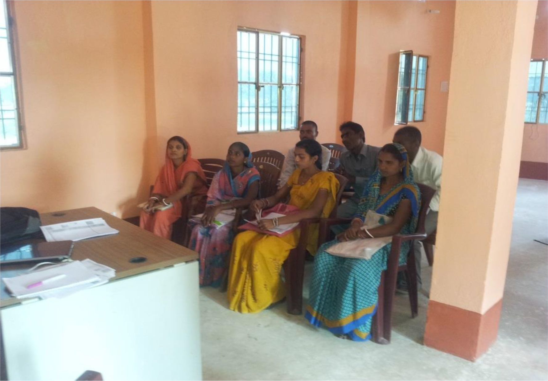 The image shows a group of villagers consisting of four women and three men with plastic bags, are seated on chairs inside a building. They are attentively looking at the front of the room. In front of them is a desk with some papers, a pen, a laptop and a bag.