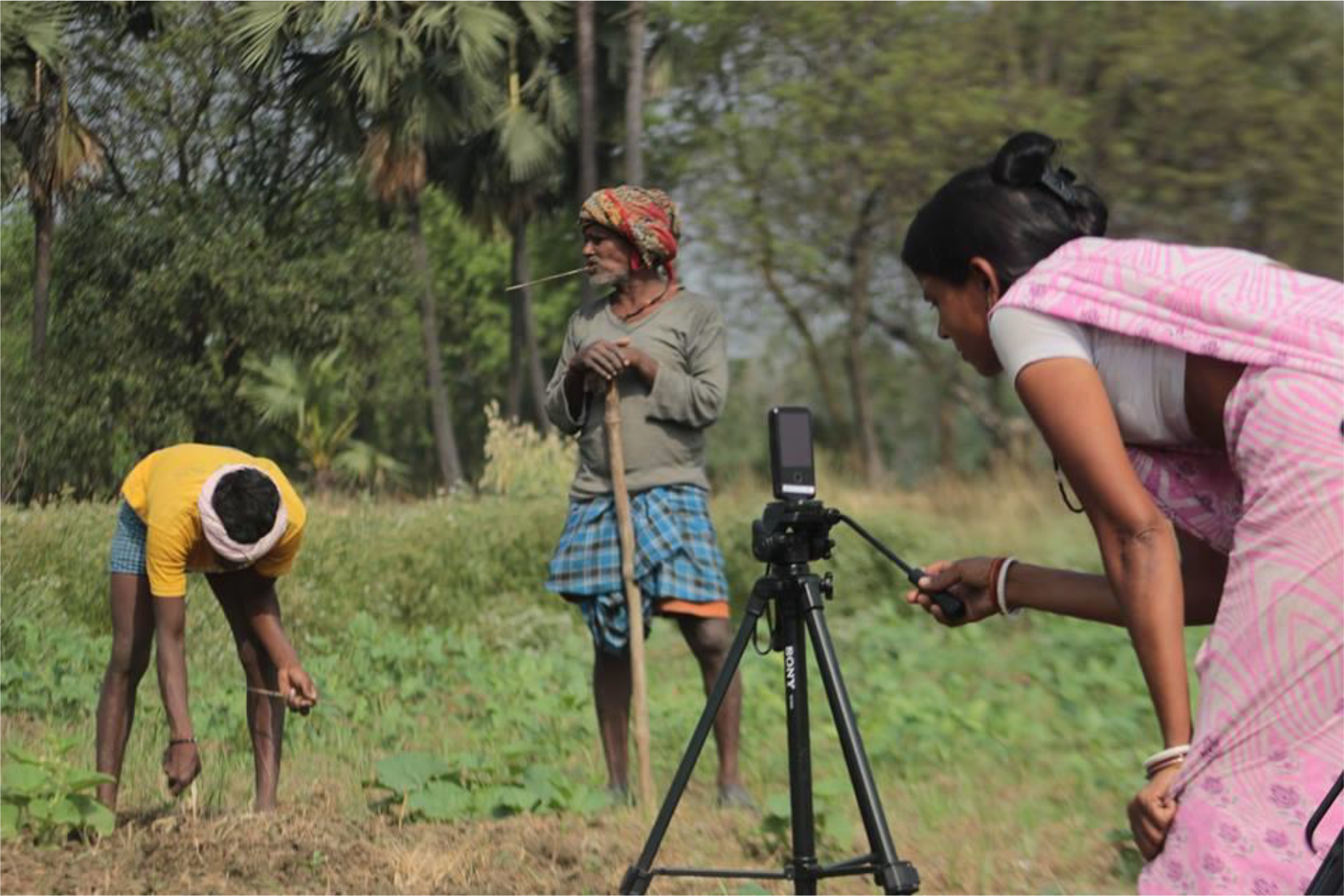 The image shows a woman shooting a video of two men working in the field.