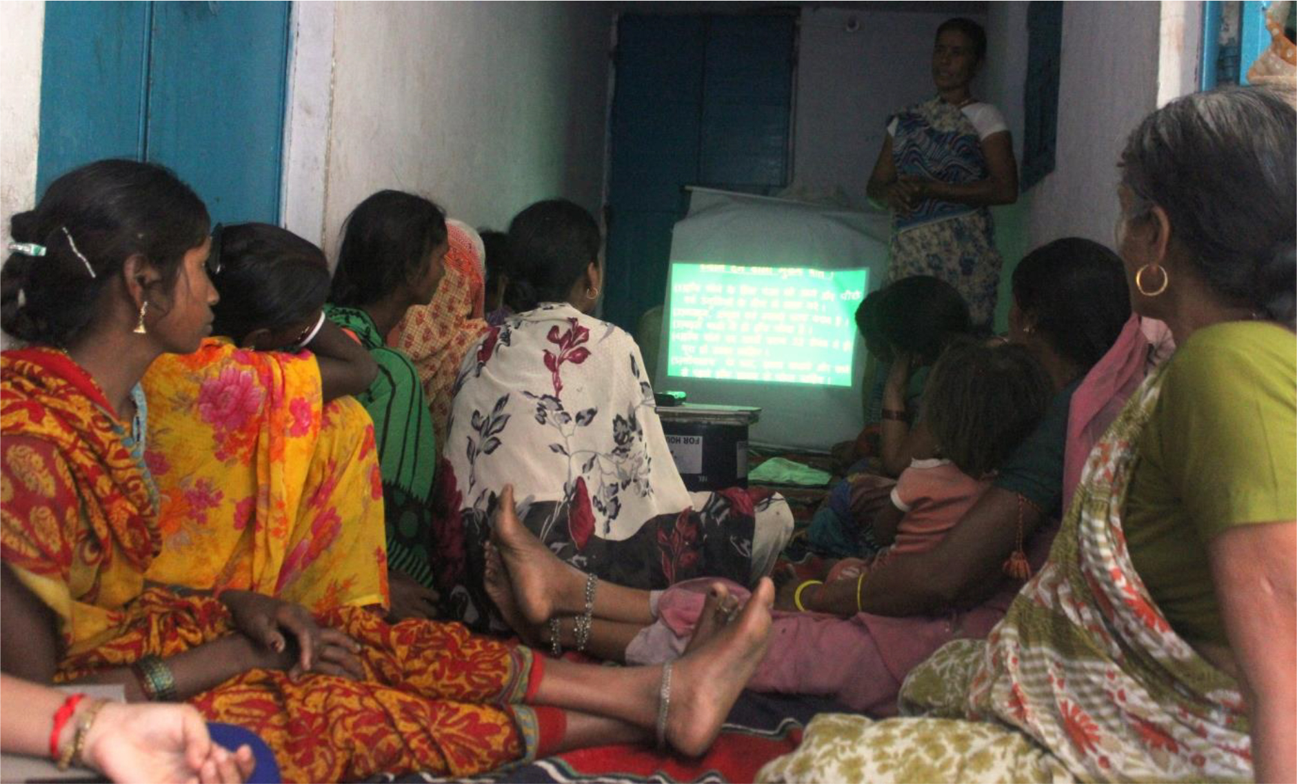 The image shows a group of village women seated on the floor attentively looking at the display from a projector.