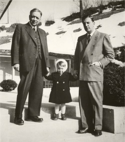 A black-and-white photograph of two men on the steps of a building with a child between them, holding both their hands. Both the men wear suits and the man on the left appears older than the man on the right. The young child wears a dark jacket and a light cap covering her head.
