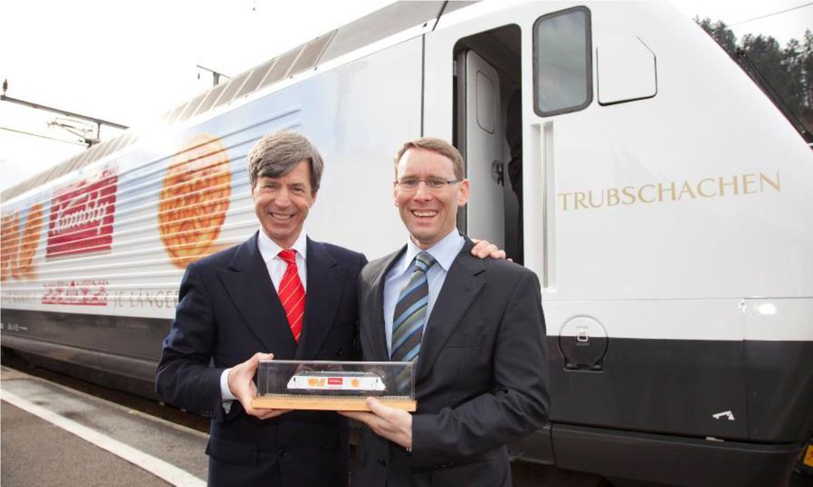 Two men in suits are seen smiling and posing in front of a train with the image of cookies on the side of the train with the Kambly logo on it. The word Trubschachen is seen on the front of the train.