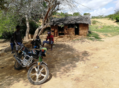 Three men sitting and one man standing under a tree in front of a tattered hut. A motorbike is parked in front of them. The men appear to be engaged in some chat.
