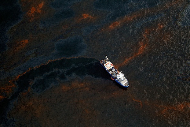 A picture shows a white boat cutting through black water which is heavily contaminated by an oil spill. The water is black with occasional orange swirls.