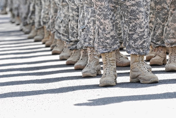 A picture shows the feet of soldiers standing at ease. They are standing uniformly in a line, such that their shadows, which fall on their right, appear to be parallel lines.
