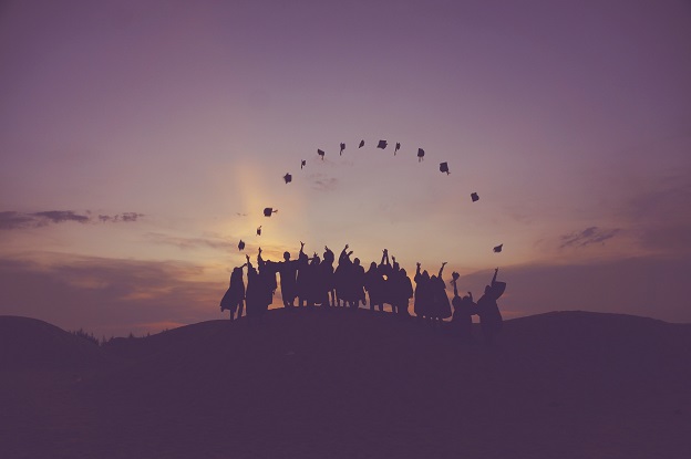 A picture shows a group of students throwing graduation caps in the air standing on a landscape of the sunset.