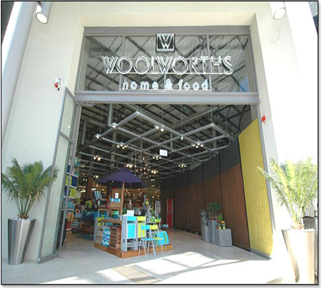 A photo shows the front view of the Woolworths Home & Food store. Text on top of the entrance reads “Woolworths home & food.” An umbrella, a few chairs, shelves, and a few plant pots are visible inside. A plant pot is on either side of the entrance.